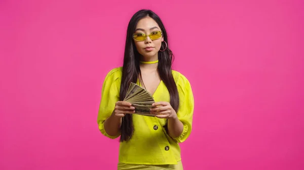 Стильна азіатська жінка в сонцезахисних окулярах тримає доларові банкноти ізольовані на рожевому — стокове фото