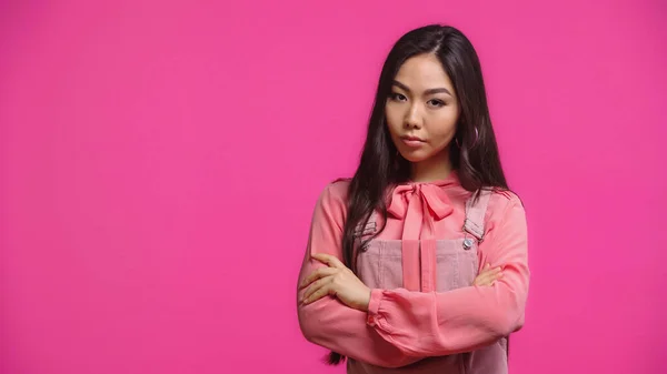 Skeptische und junge asiatische Frau blickt in die Kamera, während sie mit verschränkten Armen auf rosa — Stockfoto