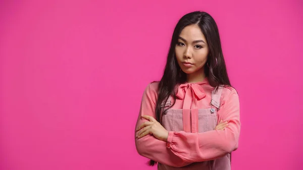 Skeptical and young asian woman looking at camera while posing with crossed arms isolated on pink — Stock Photo