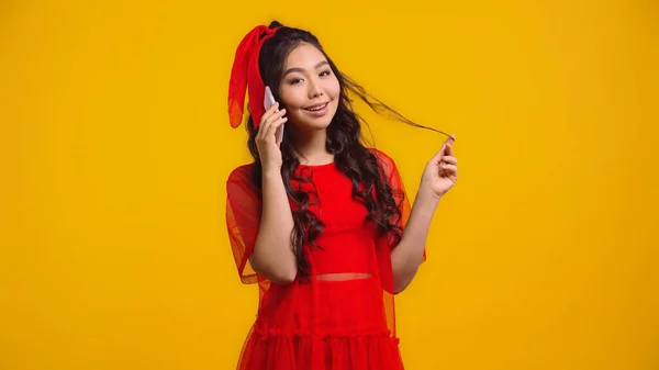 Smiling asian woman in red dress talking on smartphone while twirling hair isolated on yellow — Stock Photo