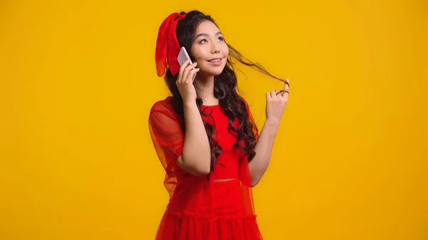 Счастливая азиатка в красном платье разговаривает на смартфоне, крутя волосы, изолированные на желтом — стоковое фото