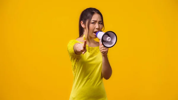 Displeased asian woman screaming in megaphone and pointing with finger isolated on yellow — Stock Photo