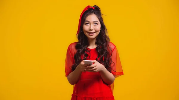 Feliz asiático mujer en rojo vestido mensajes de texto en smartphone aislado en amarillo - foto de stock