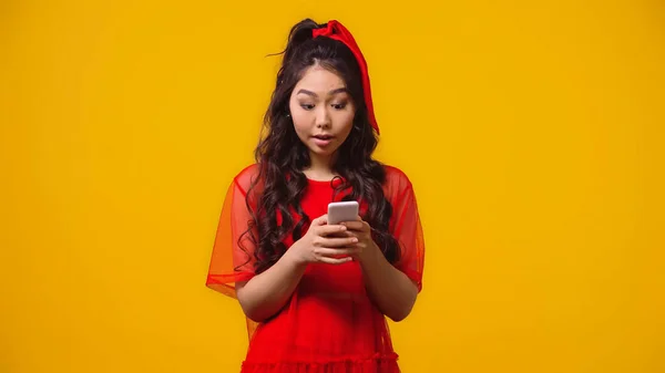 Überrascht asiatische Frau im roten Kleid SMS auf Smartphone isoliert auf gelb — Stockfoto