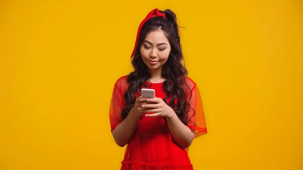Mujer asiática complacida en vestido rojo mensajes de texto en el teléfono inteligente aislado en amarillo - foto de stock