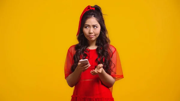Mujer asiática confusa en vestido rojo mensajes de texto en el teléfono inteligente aislado en amarillo - foto de stock