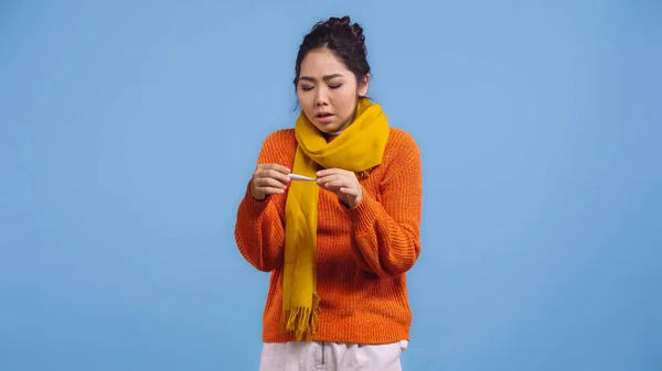 Doente mulher asiática em suéter e cachecol segurando termômetro digital isolado em azul — Fotografia de Stock