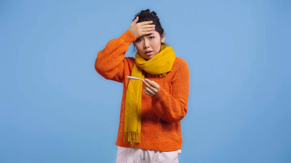 Diseased asian woman in sweater and scarf holding digital thermometer and touching forehead isolated on blue — Stock Photo
