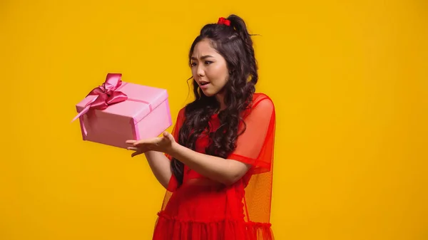 Confused asian woman holding wrapped present isolated on yellow — Stock Photo