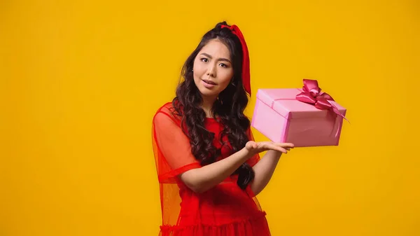 Puzzled young asian woman holding wrapped present isolated on yellow — Stock Photo