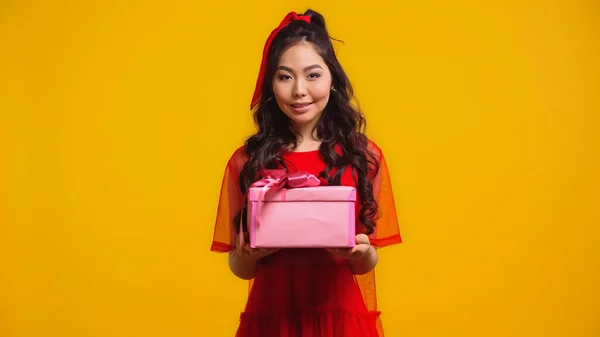 Веселая и молодая азиатская женщина держит завернутый подарок изолирован на желтый — стоковое фото