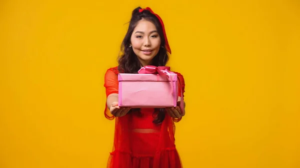 Улыбающаяся молодая азиатская женщина, держащая завернутый подарок, изолированная на желтый — стоковое фото
