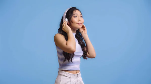 Fröhliche asiatische Frau mit drahtlosen Kopfhörern, die isoliert auf blauem Grund Musik hört — Stockfoto