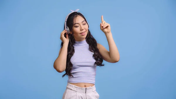 Mujer asiática feliz en auriculares inalámbricos escuchando música y señalando con el dedo aislado en azul - foto de stock