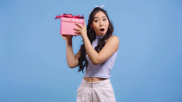 Schockierte junge asiatische Frau schüttelt umhülltes Geschenk isoliert auf blau — Stockfoto