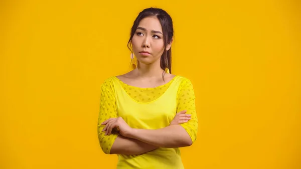 Worried and young asian woman standing with crossed arms isolated on yellow — Stock Photo