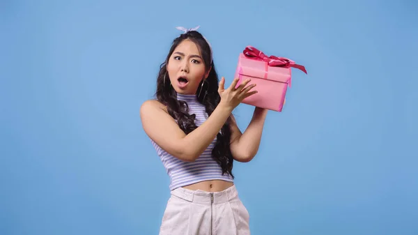Schockierte asiatische Frau schüttelt verpackte Geschenkbox isoliert auf blau — Stockfoto