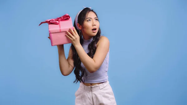 Puzzled asian woman shaking wrapped gift box isolated on blue — Stock Photo