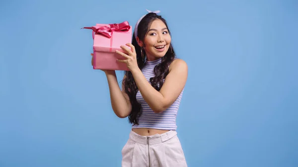 Happy young asian woman shaking wrapped present isolated on blue — Stock Photo