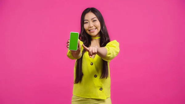 Heureux asiatique femme pointant smartphone avec écran vert isolé sur rose — Photo de stock