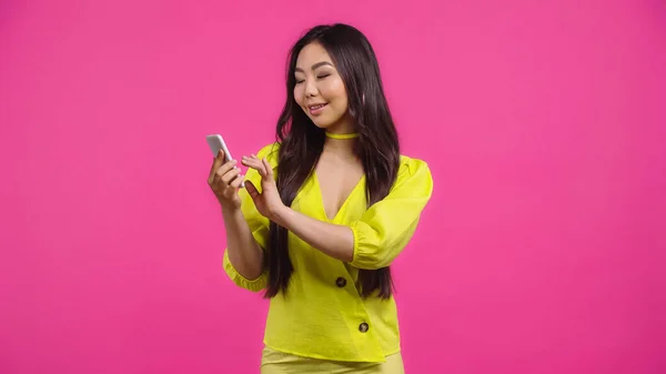 Feliz asiático mujer usando smartphone y sonriendo aislado en rosa - foto de stock