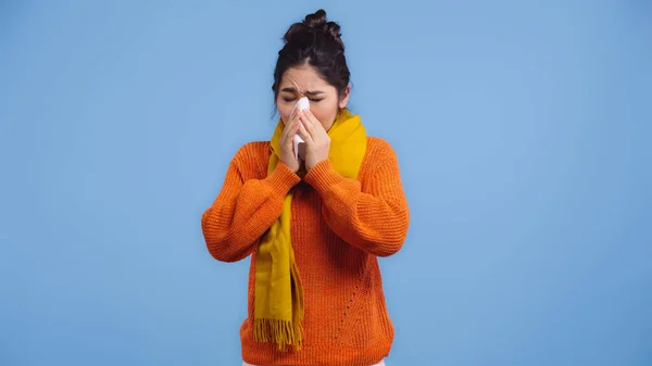 Sick asian woman in orange sweater and scarf sneezing in napkin isolated on blue — Stock Photo