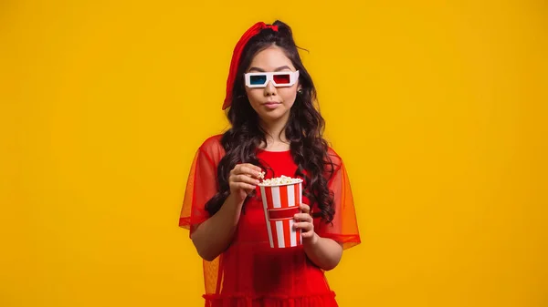 Asiático joven mujer en 3d gafas celebración palomitas de maíz y viendo película aislado en amarillo - foto de stock