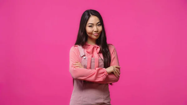 Escéptica mujer asiática de pie con los brazos cruzados y sonriendo aislado en rosa - foto de stock
