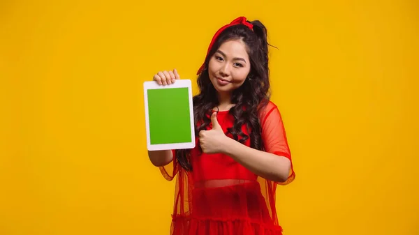 Cheerful asian woman holding digital tablet with green screen and showing like isolated on yellow — Stock Photo