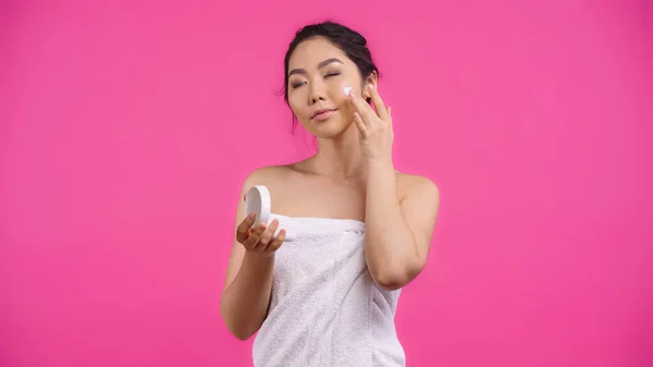 Asian young woman in towel applying face cream isolated on pink — Stock Photo