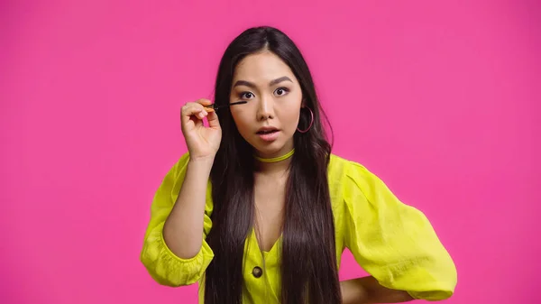 Fokussierte asiatische Frau, die Mascara aufträgt und isoliert auf rosa lächelt — Stockfoto