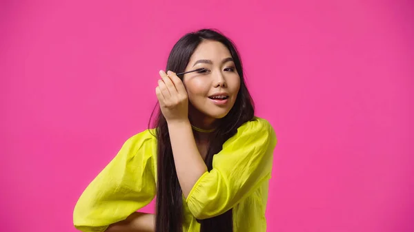 Alegre asiático mujer aplicando rímel y sonriendo aislado en rosa - foto de stock