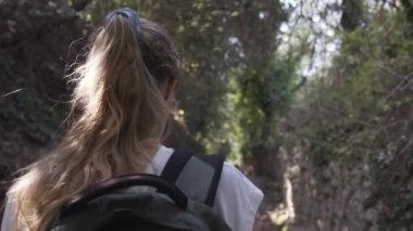 Sarışın yürüyüşçü kayalık bir yolda yürüyor. Turist zeytin ağaçları arasında doğayı keşfeder. Arkadan bak, açık havada yaşa, sırt çantanı topla, sarışın at kuyruğu. Yavaş çekimde 4k video.