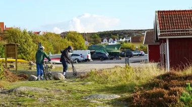 Grebbestad, İsveç - 05 10 2022: genç çift bisiklete biniyor. İsveçliler iskele yakınlarında bisiklet sürerler. Tekneler ve arabalar kırmızı ahşap evlerin arasına park edilmiş. Spor ve açık hava hayatı.