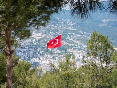 Alanya şehrinin üzerinde yeşil çam ağaçları ve uzak şehir manzarası üzerinde dalgalanan bir Türk bayrağı