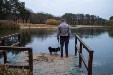 Erkek köpek sahibi ve arkadaşı göl kenarındaki ahşap iskelede duruyorlar ve sonbahar mevsiminde yürüyüşleri sırasında manzaranın tadını çıkarıyorlar. İnsan ve evcil hayvan konsepti.