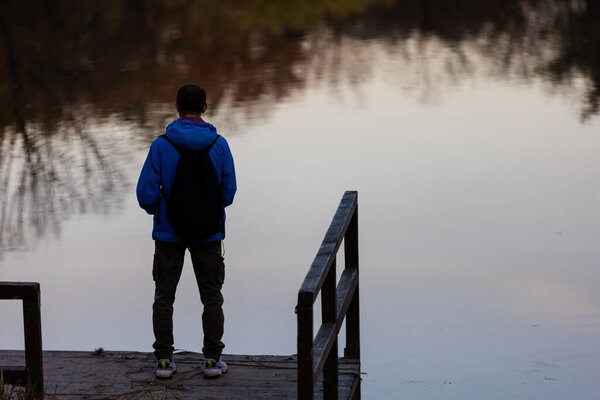 Young man with backpack standing alone on wooden footbridge and staring at lake. Peaceful atmosphere in nature. Enjoying fresh air in autumn evening. Back view.