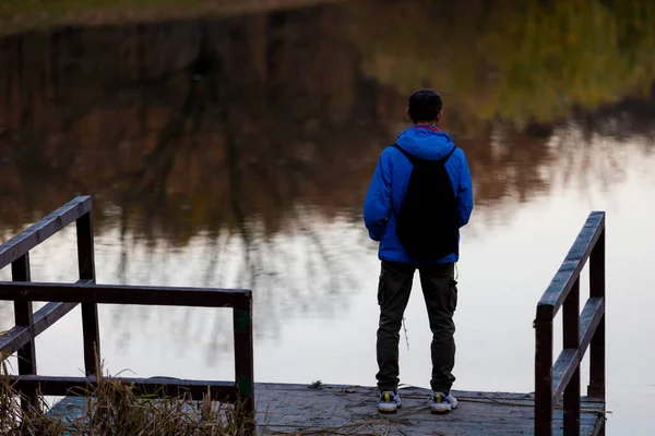 stock image Young man with backpack standing alone on wooden footbridge and staring at lake. Peaceful atmosphere in nature. Enjoying fresh air in autumn evening. Back view.