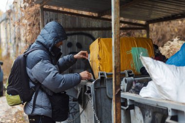 Ukrayna 'nın kentsel caddesindeki çöplükte yiyecek arayan fakir ve aç evsiz bir adam. Modern toplumun sosyal sorunları. Sokak serserilerinin yaşam tarzı, sokaklarda yaşamak..