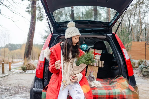 stock image Happy Asian girl in white sweater sitting with Christmas bunny and gift boxes in car trunk. 