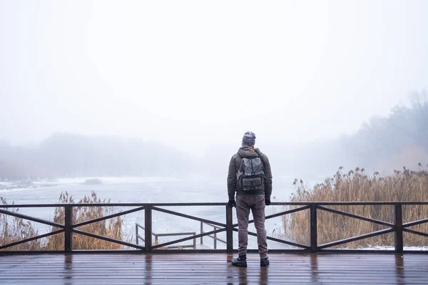 Young man in fog standing alone on wooden footbridge and staring at lake. Hooded guy. Peaceful atmosphere in nature. Enjoying fresh air in winter. Back view.