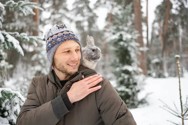 Close portrait of a man with a rabbit. A gray fluffy bunny sits on shoulder of his owner in winter snowy forest. Travel with pet.