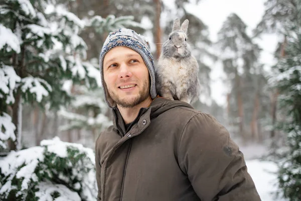 Close portrait of a man with a rabbit. A gray fluffy bunny sits on shoulder of his owner in winter snowy forest. Travel with pet.