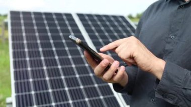 a man holds a smartphone near solar panels on a plot near the house.