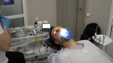 Bir erkek dişçi, sandalyede yatay olarak yatan bir adamın dişinde delik açma sürecinde. Mavi üniformalı ve maskeli ortodontist.