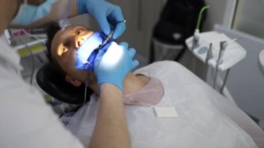 Bir erkek dişçi, sandalyede yatay olarak yatan bir adamın dişinde delik açma sürecinde. Mavi üniformalı ve maskeli ortodontist.