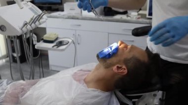 Bir erkek dişçi, sandalyede yatay olarak yatan bir adamın dişinde delik açma sürecinde. Mavi üniformalı ve maskeli ortodontist.