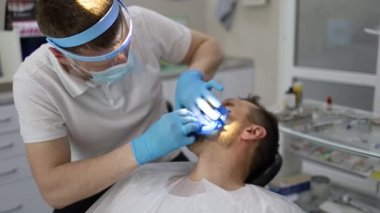 Bir erkek dişçi, sandalyede yatay olarak yatan bir adamın dişinde delik açma sürecinde. Mavi üniformalı ve maskeli ortodontist.