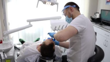 Bir erkek dişçi, sandalyede yatay olarak yatan bir adamın dişinde delik açma sürecinde. Mavi üniformalı ve maskeli ortodontist.