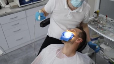 Bir erkek dişçi, sandalyede yatay olarak yatan bir adamın dişinde delik açma sürecinde. Mavi üniformalı ve maskeli ortodontist.